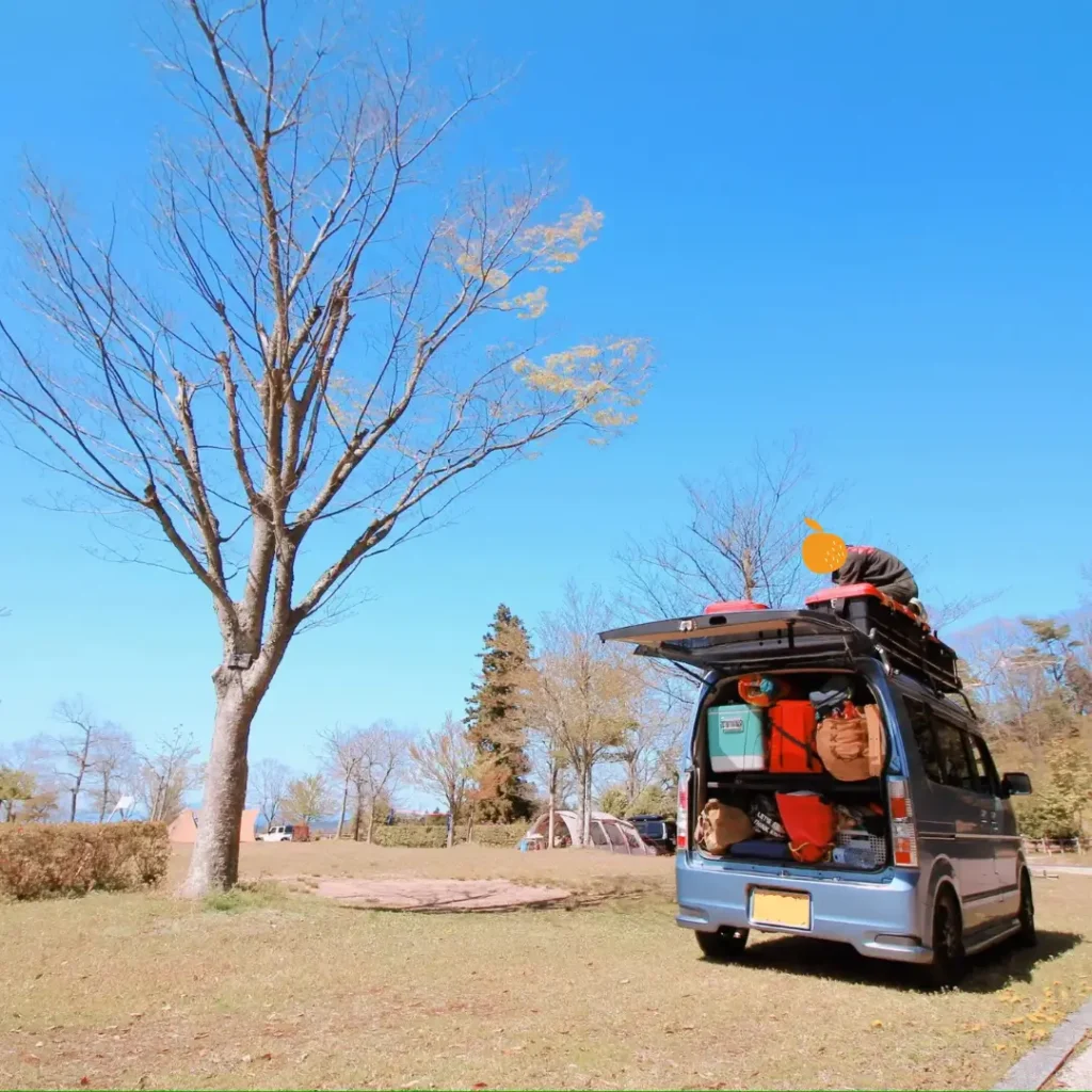 晴天のキャンプ場サイトに駐車し、バックドアを開けてキャンプ道具が満載状態のスズキ・エブリイワゴン。ルーフキャリアにも荷物を積み、使い慣れた「こなれ感」が漂うアウトドア風景の写真。