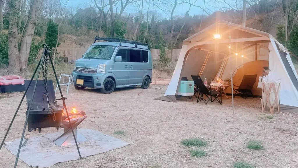 キャンプサイトで焚き火と大型テントの横に佇むスズキ・エブリイワゴン。軽自動車という選択が、豊かなレジャーと家計の余裕を両立させていることを示すライフスタイル写真。